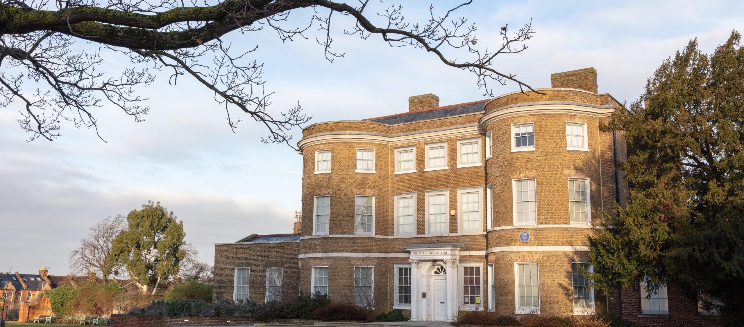 Image of William Morris Gallery's exterior. The image is taken at an angle and we can see a tree branch in the foreground, which frames the image.