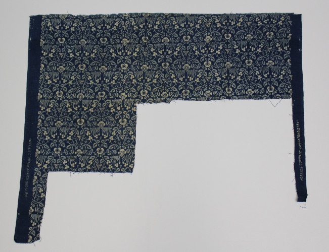 Textile sample with large sections cut out from the fabric. Design features small flowers, leaves and tufts of grass all in pale blue and white on indigo cloth.